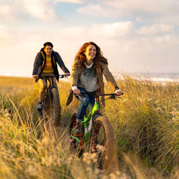 young couple bike riding in a long grass path near the beach
